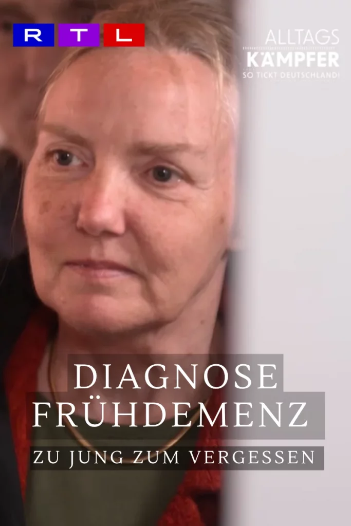 Auf dem Bild sieht man eine ältere Dame mit neutralem bis leicht traurigem Blick. Sie hat einen kleinen Leberfleck unter dem linken Auge, trägt ein rotes Jackett, darunter ein grünes Oberteil und eine goldene Halskette.