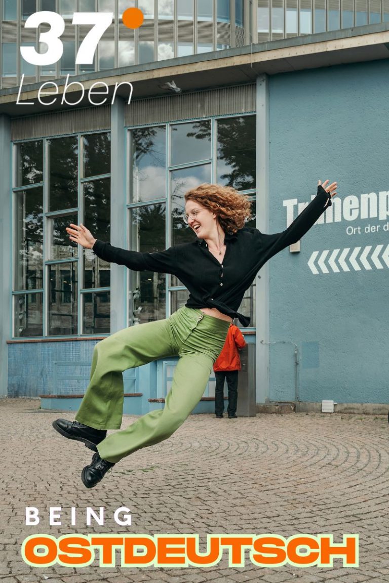 Foto einer jungen Frau mit grüner Hose und langen, rötlich gelockten Haaren. Sie springt nach rechts hoch und ist fröhlich. Hinter ihr ist ein Gebäude zu sehen, der Tränenpalast. Das Logo 37 Grad Leben und der Titel der Sendung: Being Ostdeutsch ist am unteren Bildrand zu sehen.